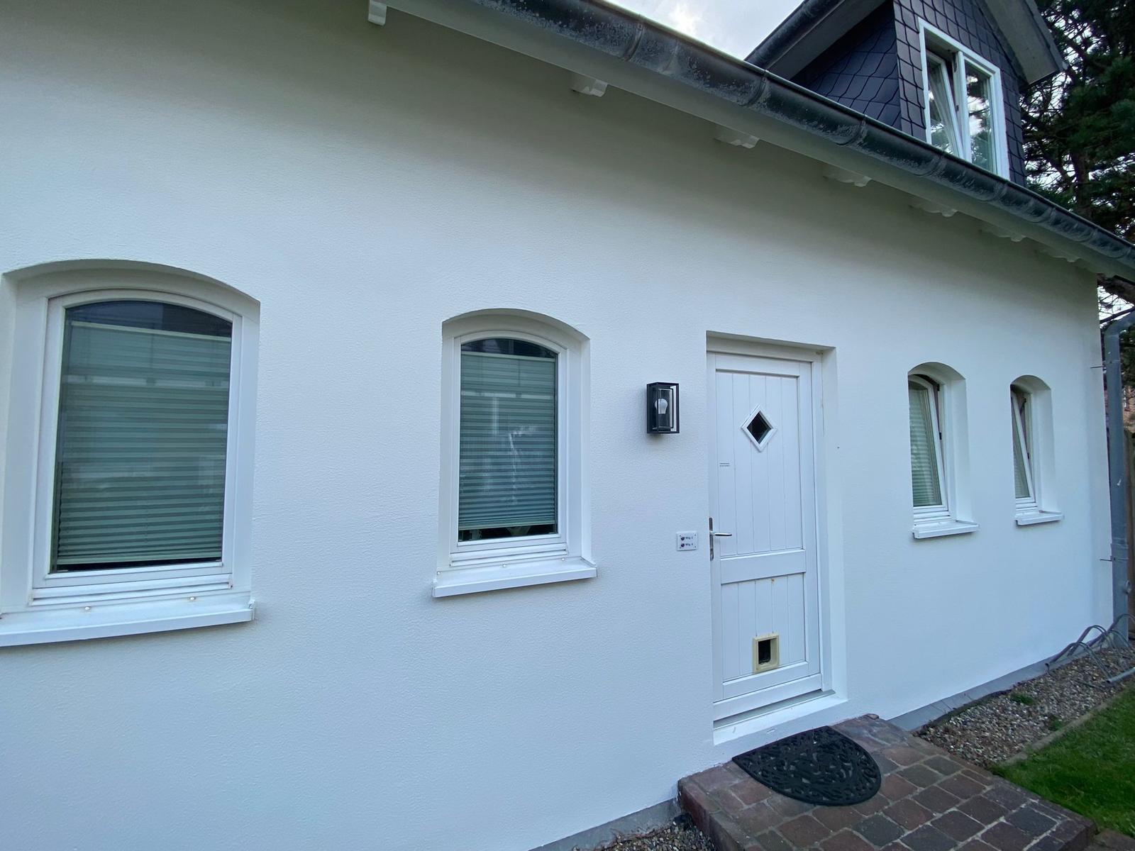 Weiße Fassade mit Eingangstür und Fenstern. Dach mit Schieferziegeln.