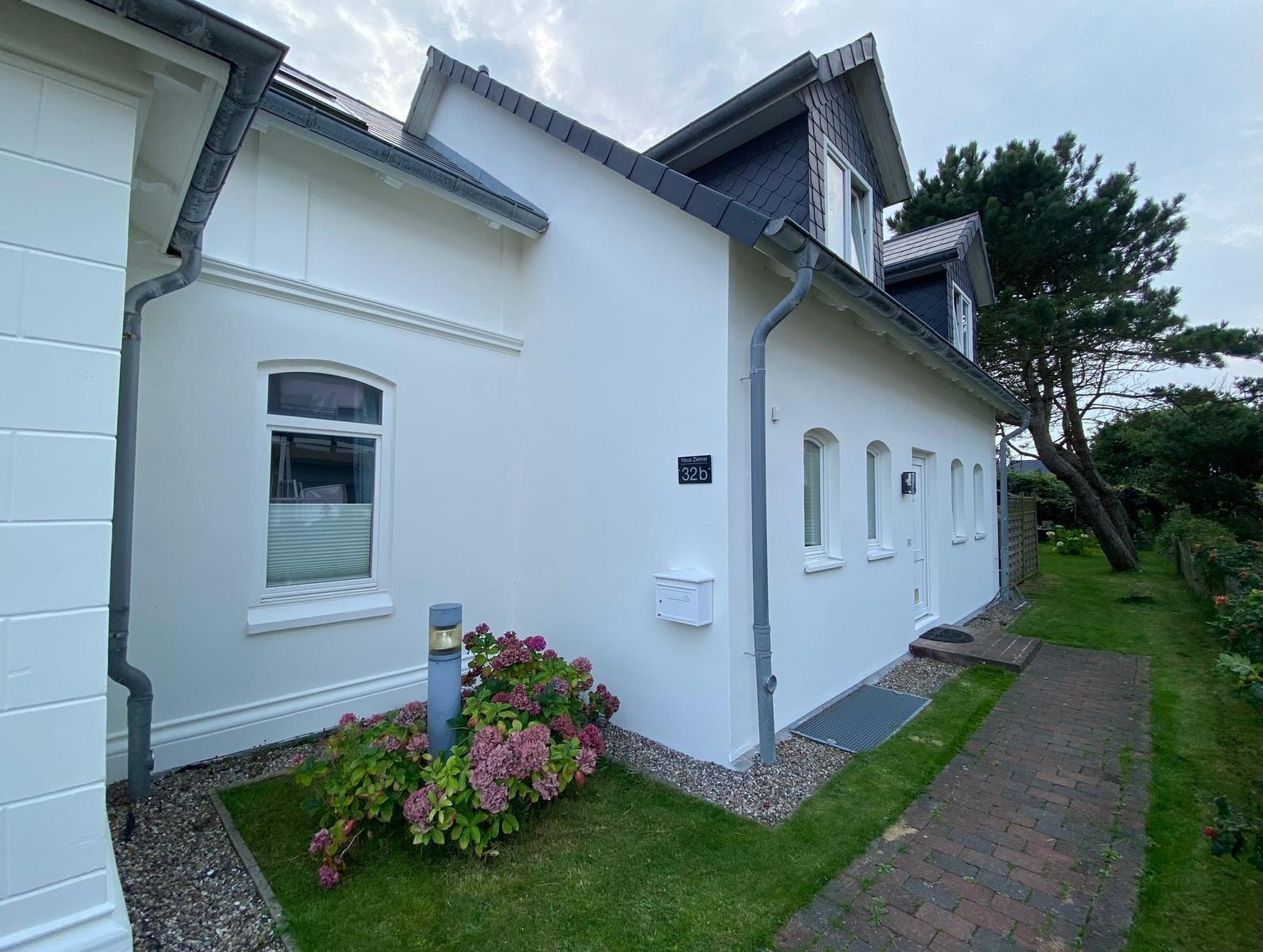 Weiße Ferienhausfassade mit Gartenweg und Blumenbeet. Dachgauben und Eingangstür sichtbar.