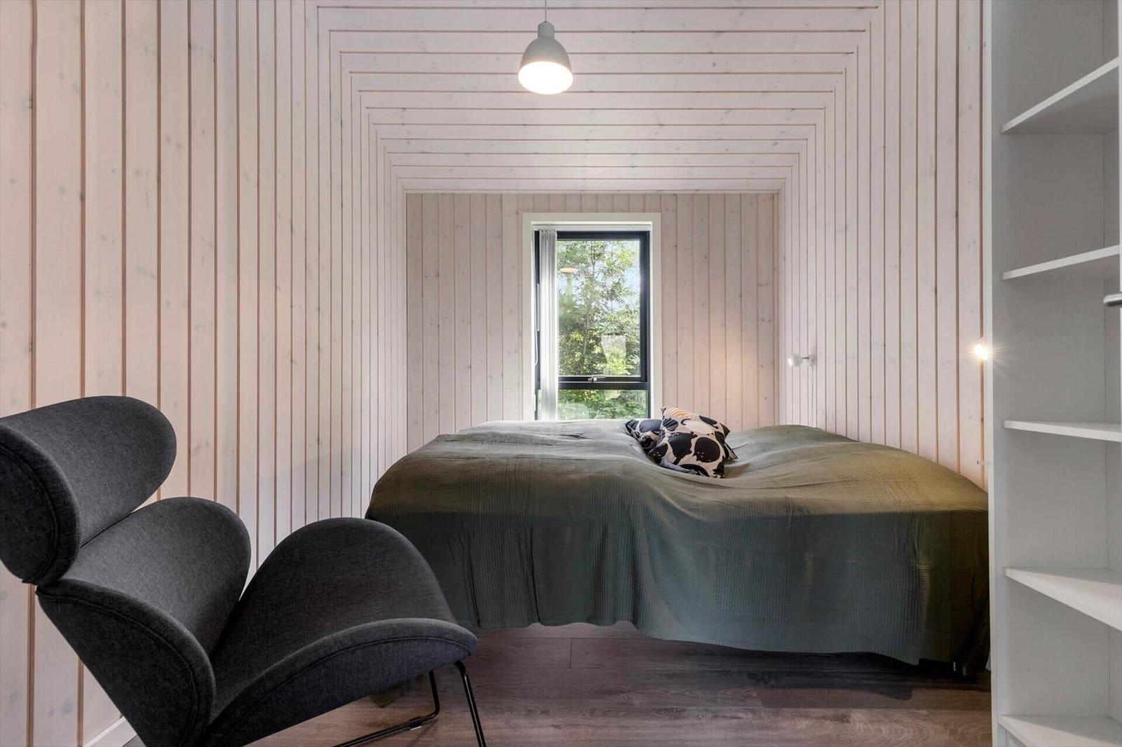 Schlafzimmer mit Bett, Fenster und grauer Sitzgelegenheit. Holzwandverkleidung und Regal.