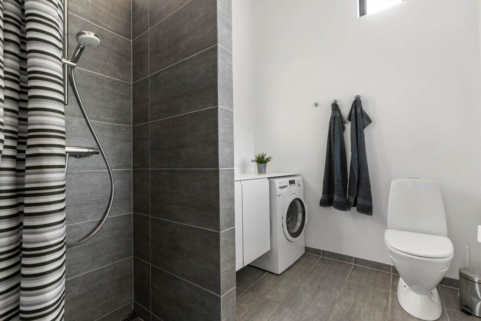 Badezimmer mit Dusche, Waschmaschine und Toilette. Graue Fliesen und weiße Wände.