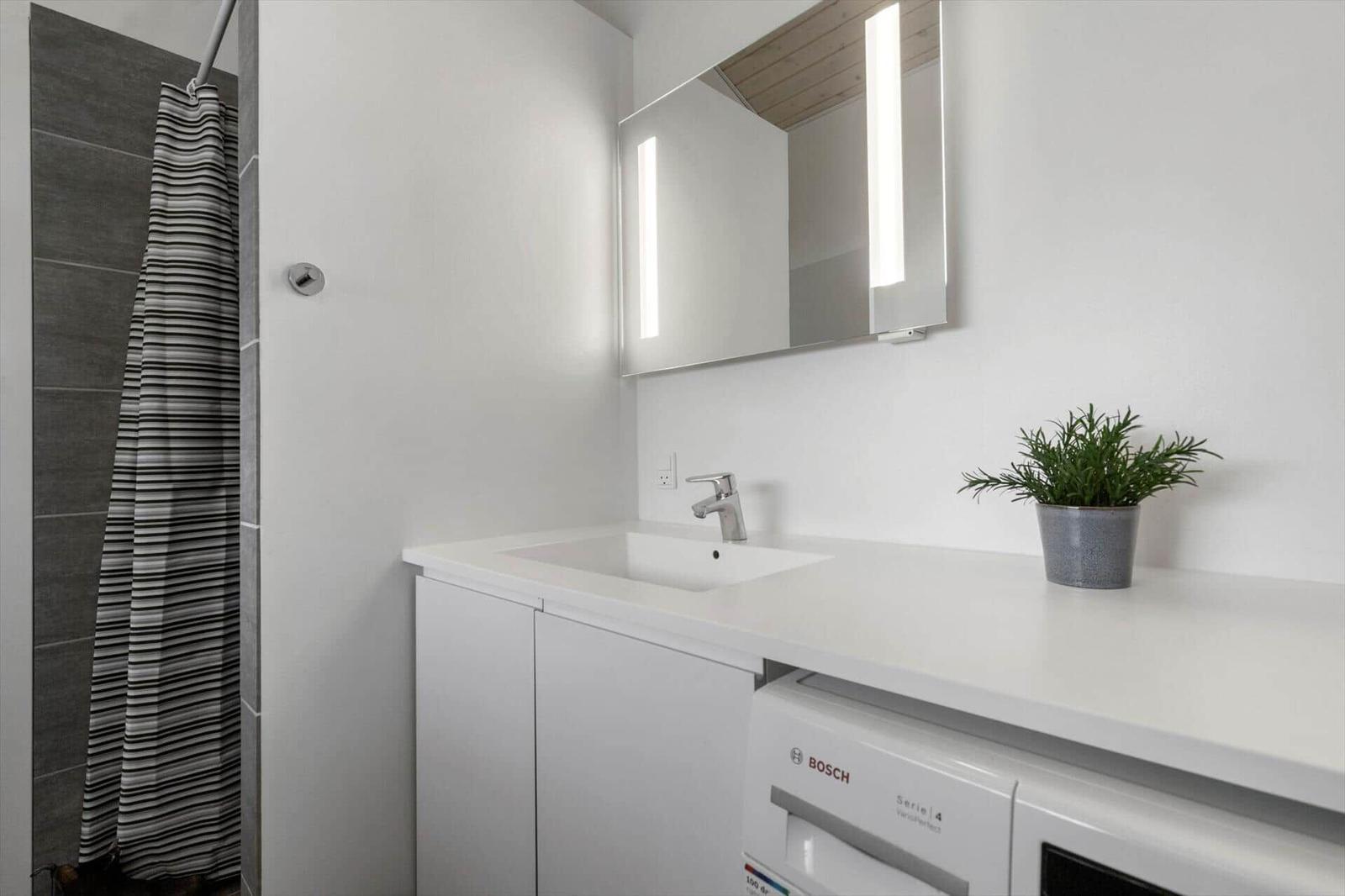 Badezimmer mit Waschbecken, Spiegel und Dusche. Unter dem Waschtisch steht eine Waschmaschine.