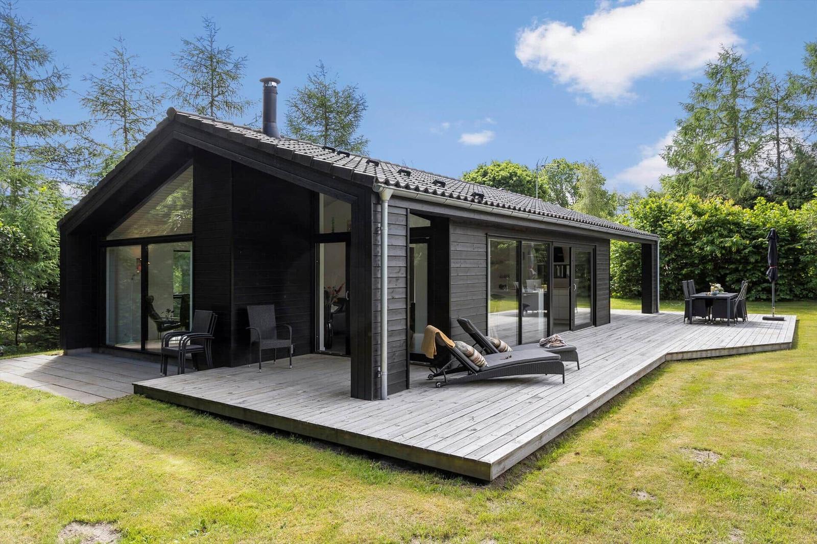 Moderne Villa mit Holzterrasse, Liegestühlen und Garten. Große Glasfronten und dunkle Holzfassade.