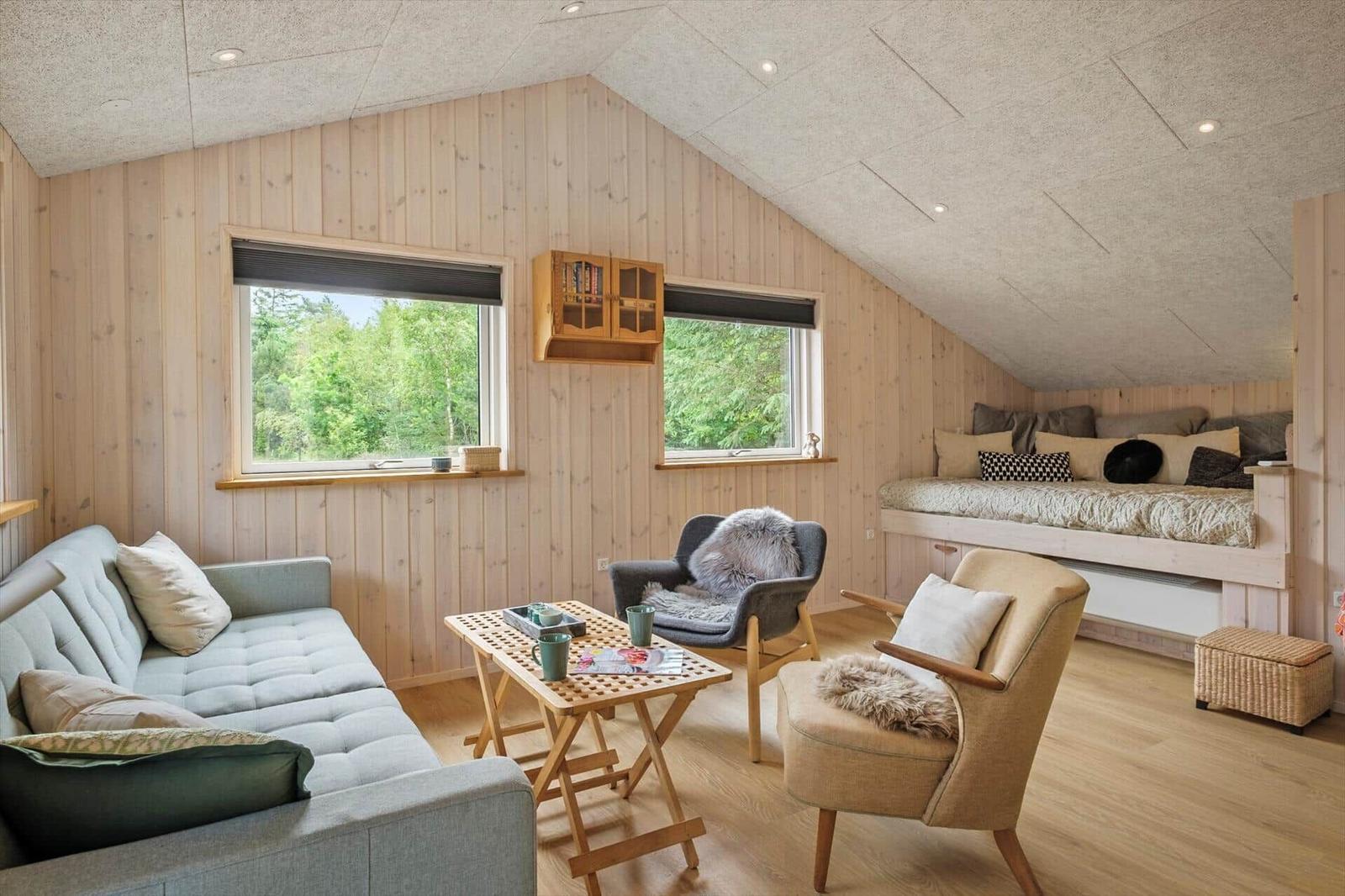 Wohnraum mit Sofa, zwei Sesseln und Schlafcouch unter Dachschrägen. Holzwände und Fenster mit Blick in die Natur.