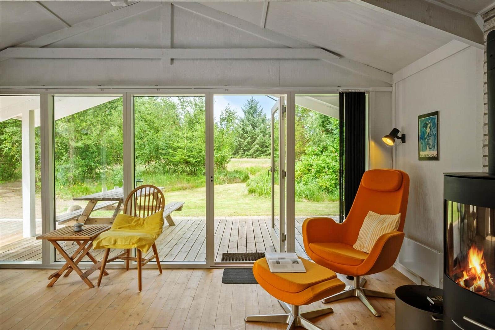 Ein Raum mit orangefarbenem Sessel, Kamin und großem Fenster zum Garten.