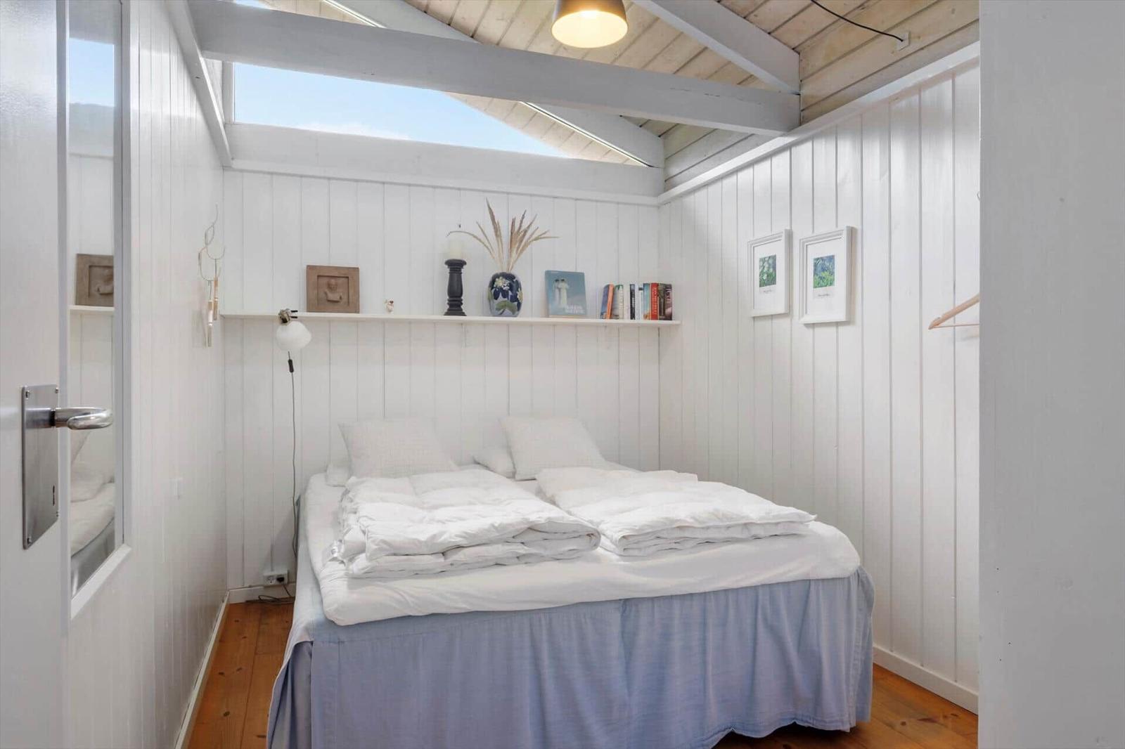 Schlafzimmer mit Doppelbett, weißen Wänden und Holzboden. Deckenfenster und Regal mit Dekor.