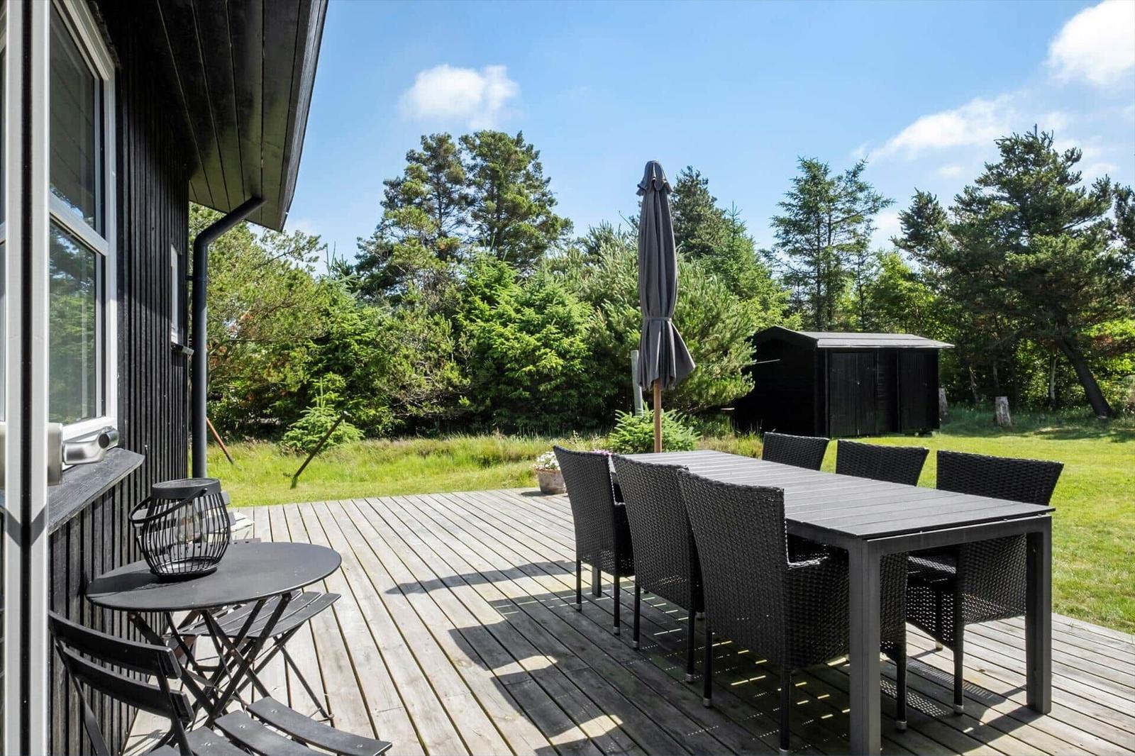 Holzterrasse mit Tisch, Stühlen und Sonnenschirm. Grünfläche und Bäume im Hintergrund.