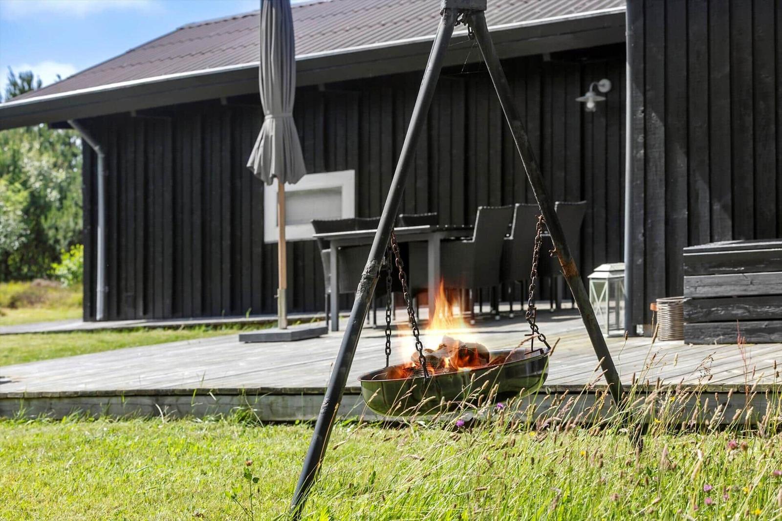 Feuerkorb auf Terrasse vor dunklem Holzhaus mit Grillplatz und Garten.