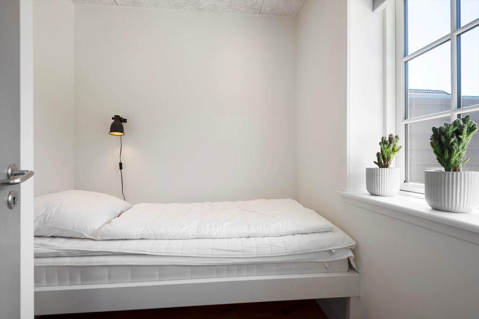 Schlafzimmer mit weißem Bett, Wandlampe und Pflanzen am Fenster.
