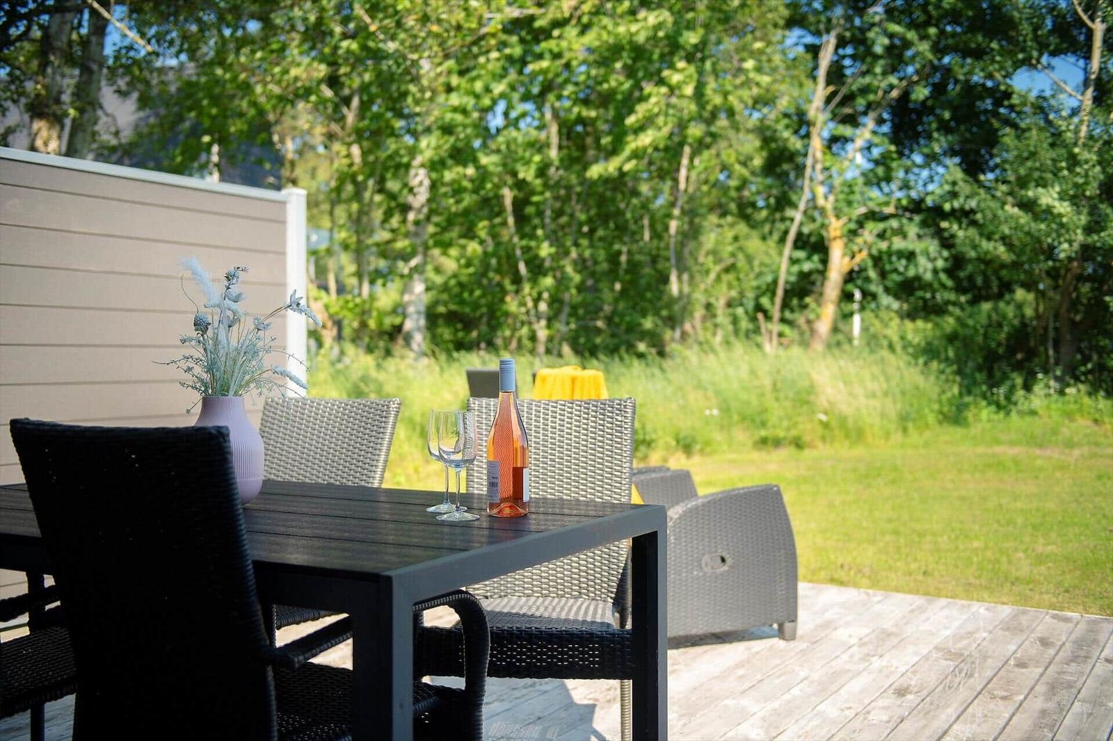 Terrasse mit Tisch, Stühlen und Weinflasche vor grünem Hintergrund.