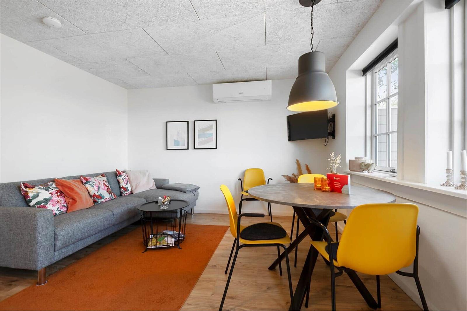 Wohn- und Essbereich mit grauem Sofa, orangefarbenem Teppich und gelben Stühlen um einen runden Tisch.