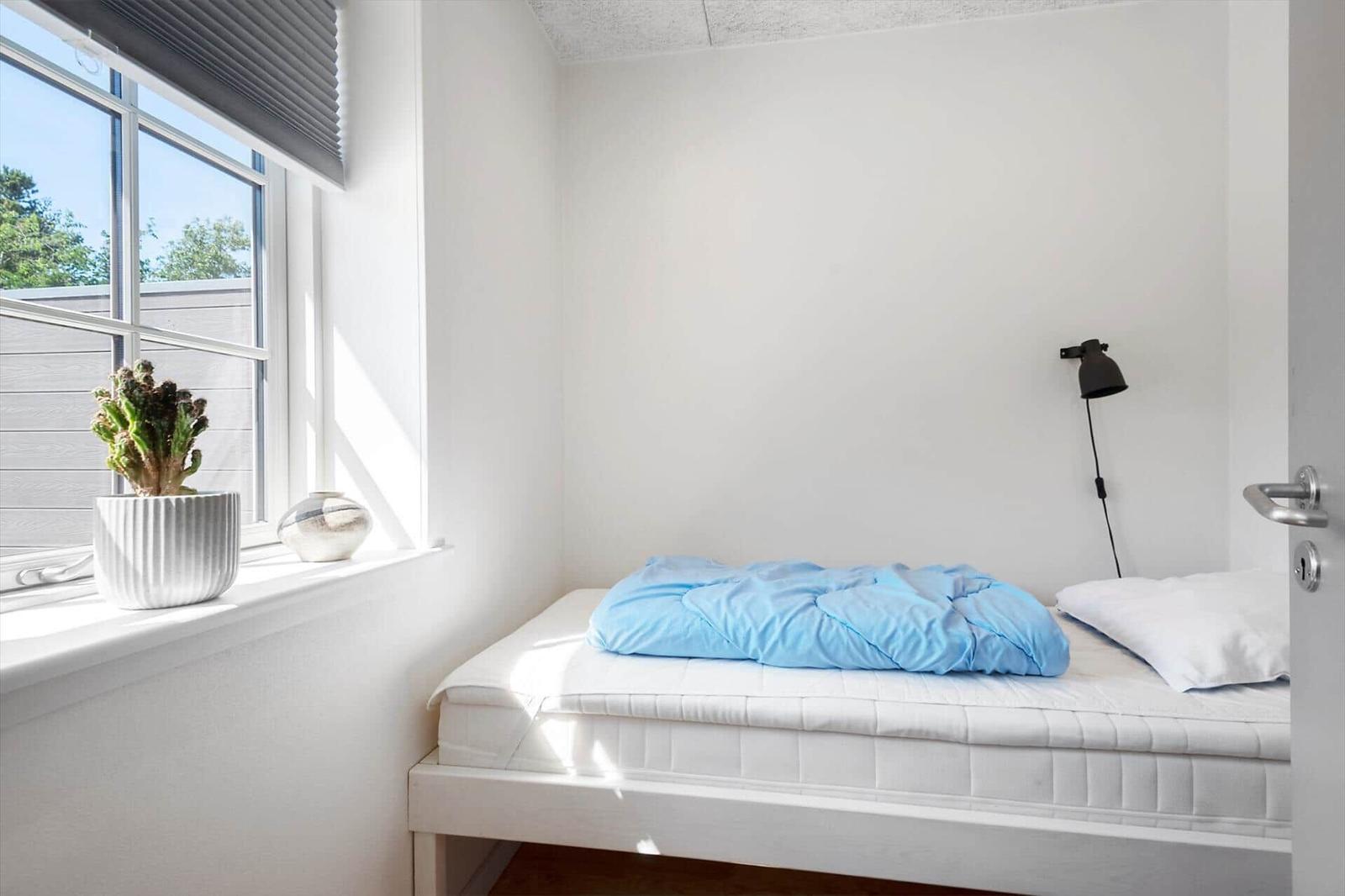 Schlafzimmer mit Bett, Fenster und Pflanze. Wandlampe und weißer Rahmen.