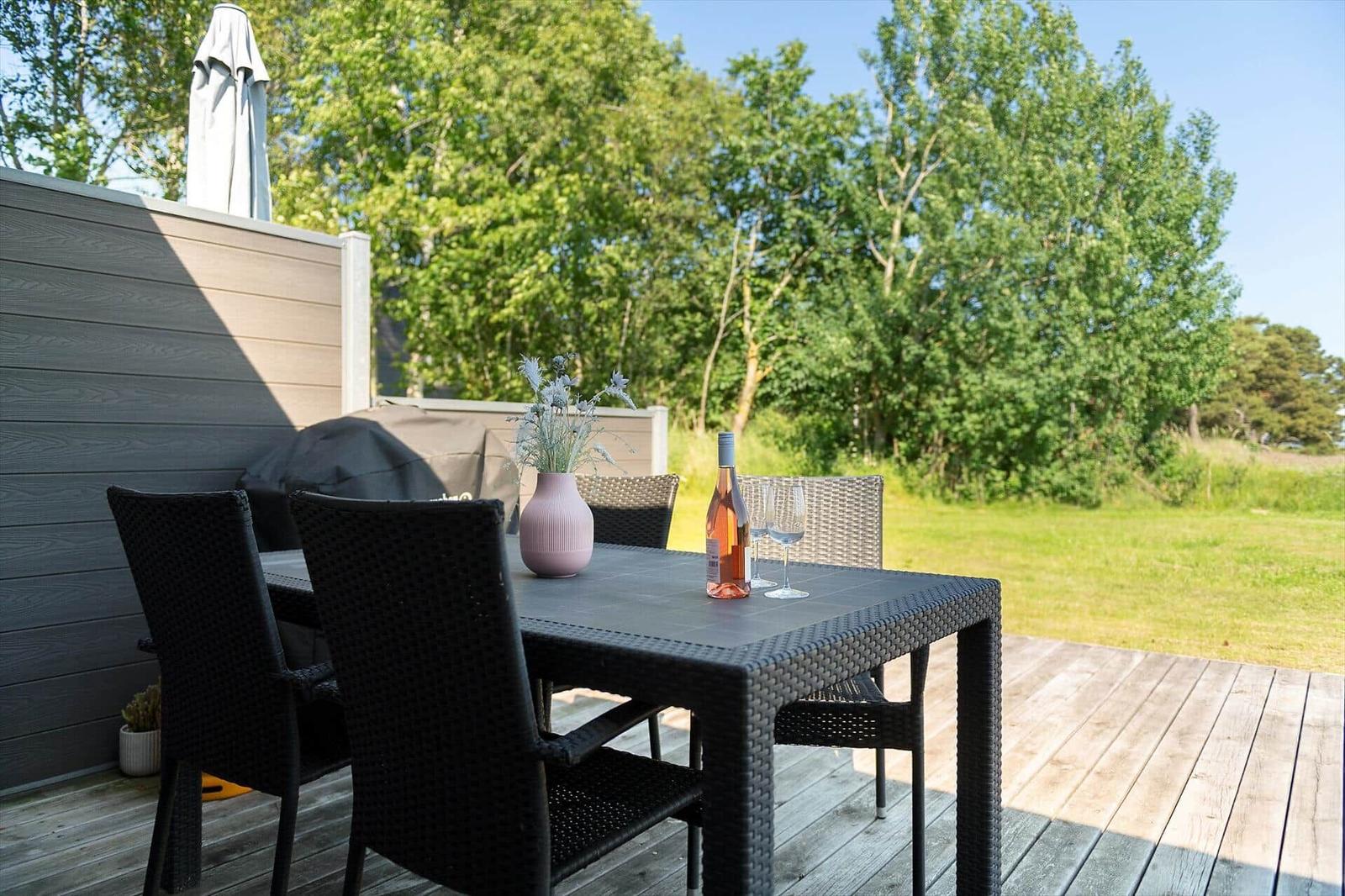 Terrasse mit Tisch, Stühlen, Wein und Blumenvase. Hintergrund: Grünfläche und Bäume.