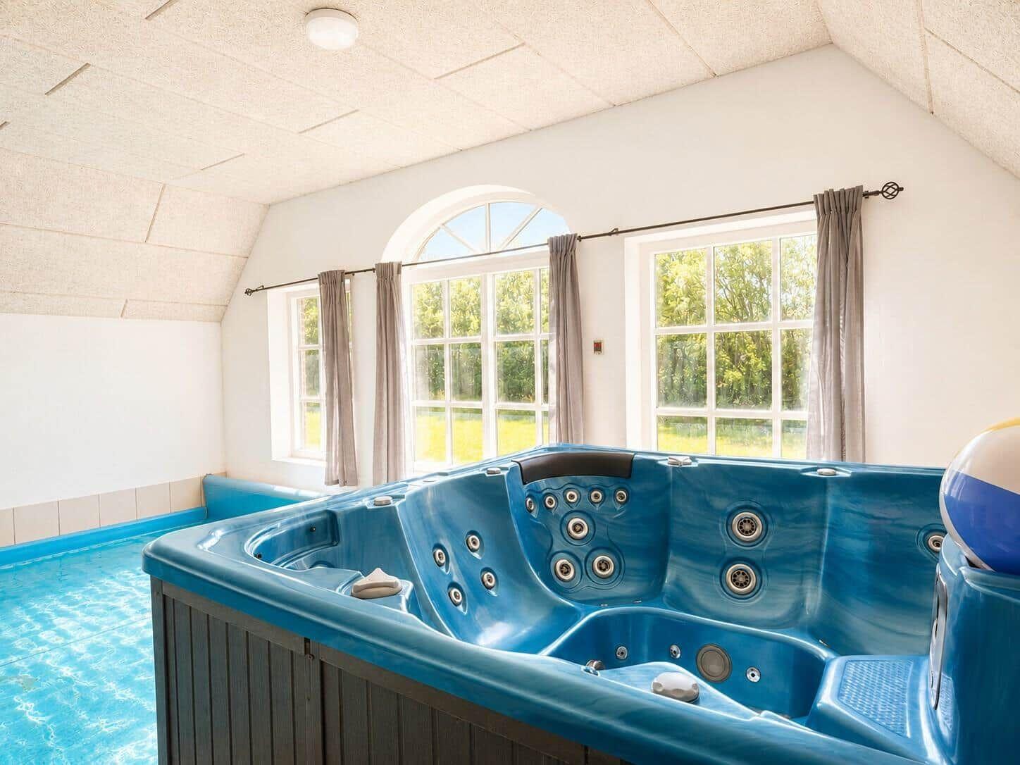 Ein blauer Whirlpool mit mehreren Düsen ist in einem Raum mit großen Fenstern und einer Dachschräge platziert.
