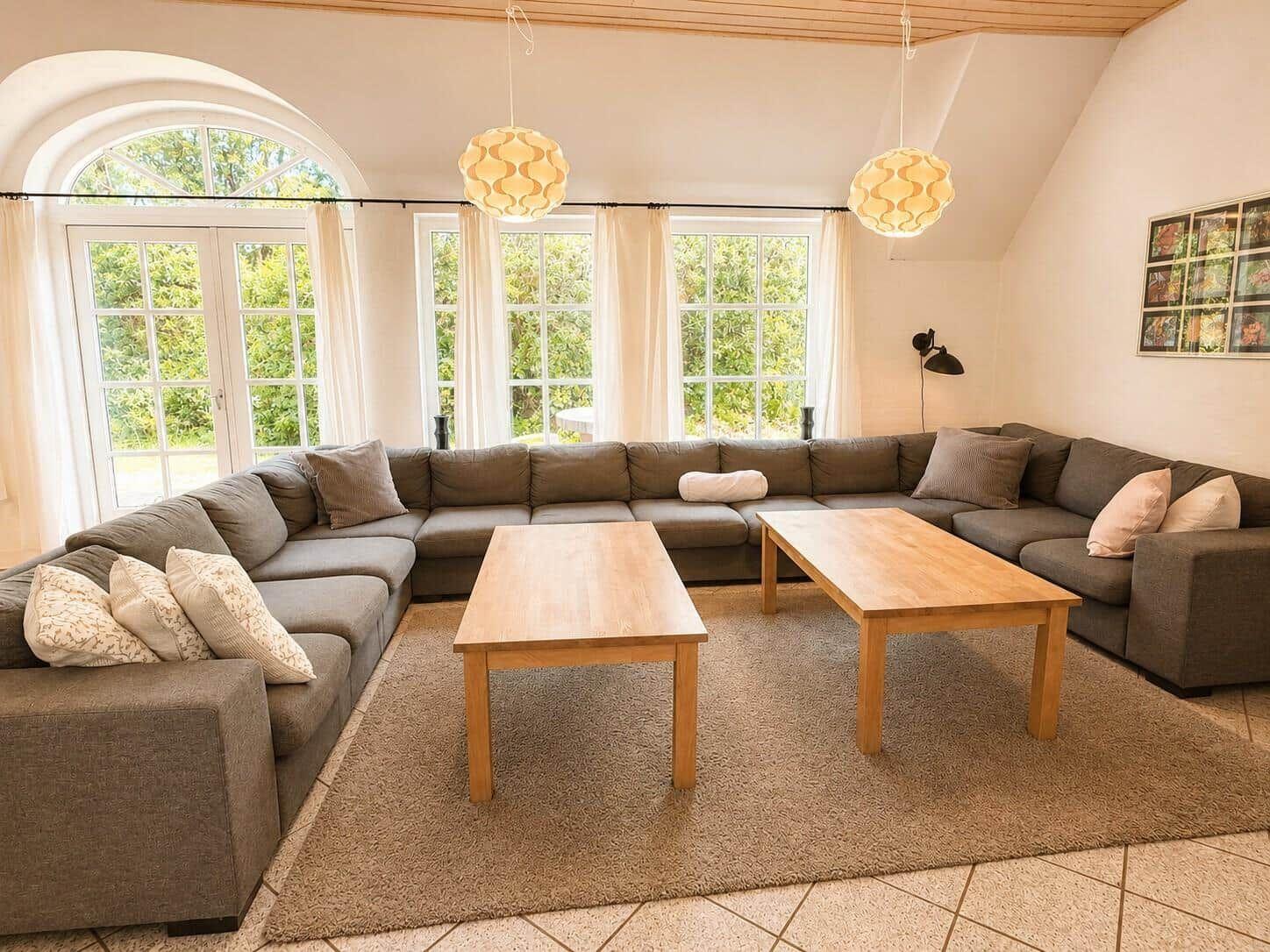 Großer Wohnbereich mit L-förmigem Sofa, zwei Holztischen und großen Fenstern.
