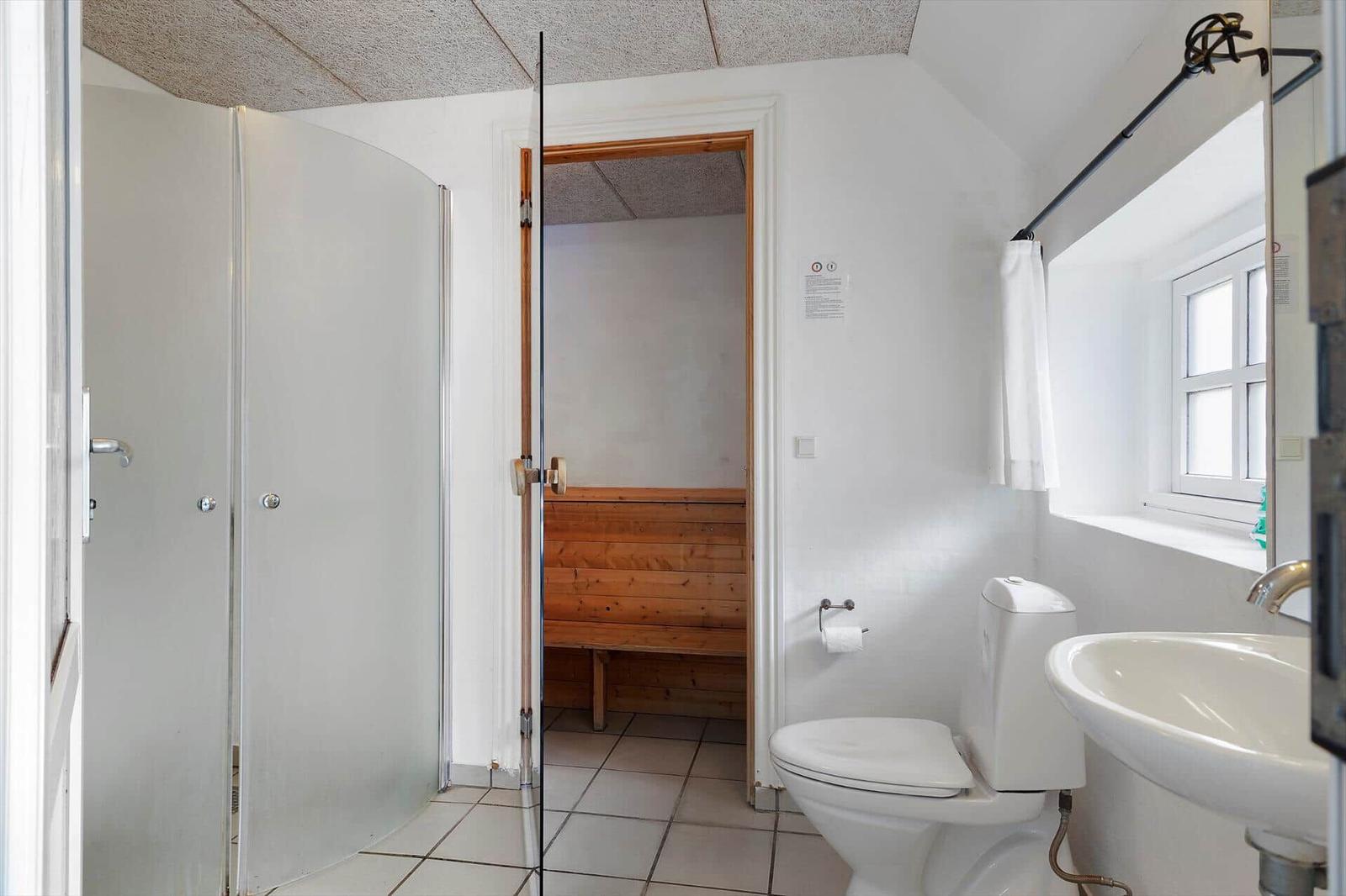 Badezimmer mit Dusche, Toilette und Waschbecken. Durchgang zur Sauna mit Holzbank.