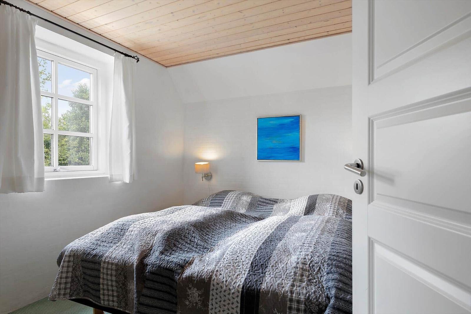 Schlafzimmer mit Bett, Fenster und Wandbild. Holzdecke und weiße Wände.