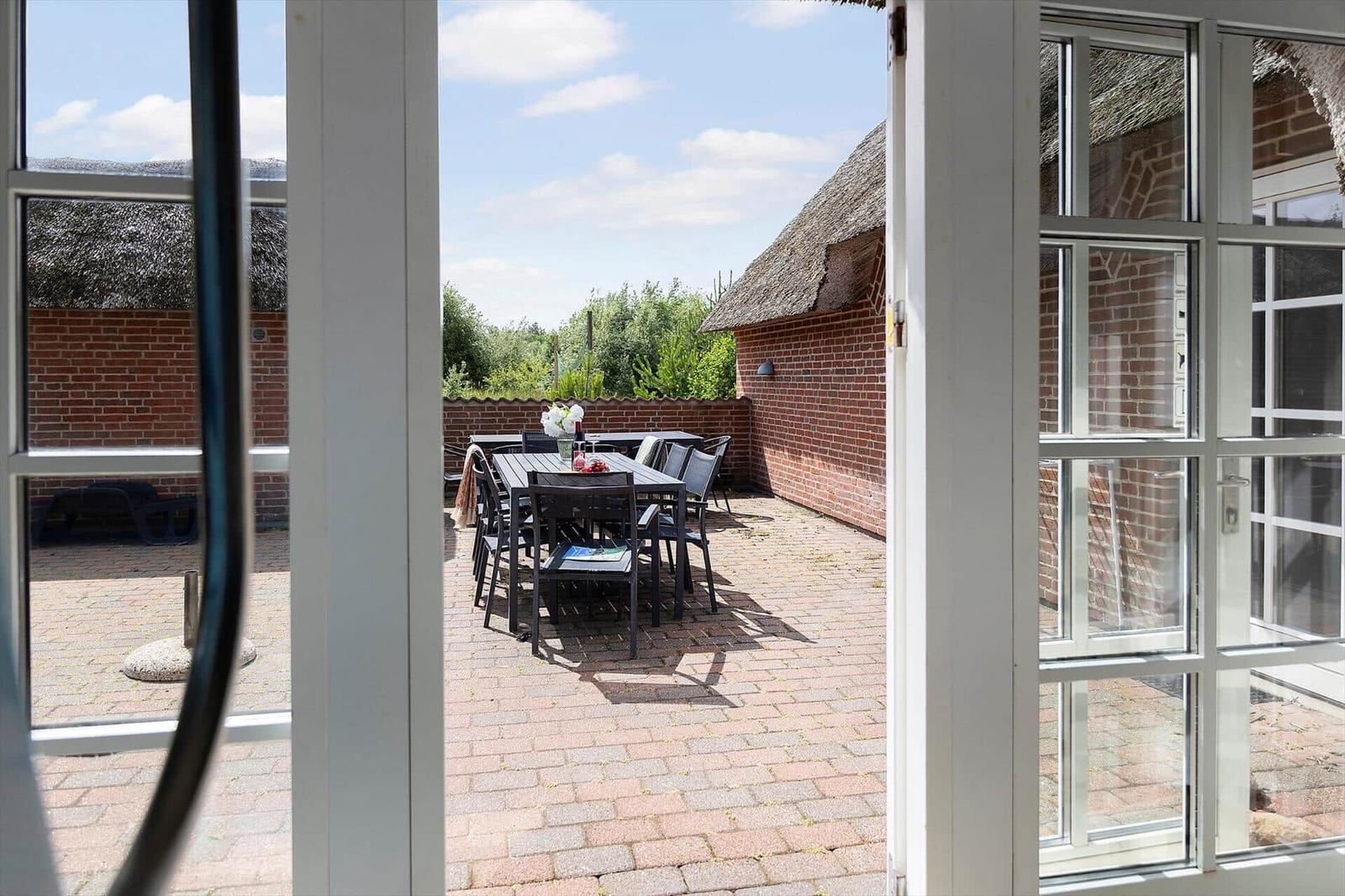 Durch die Tür blickt man auf eine Terrasse mit Tisch und Stühlen. Im Hintergrund ist ein Backsteinhaus mit Strohdach.