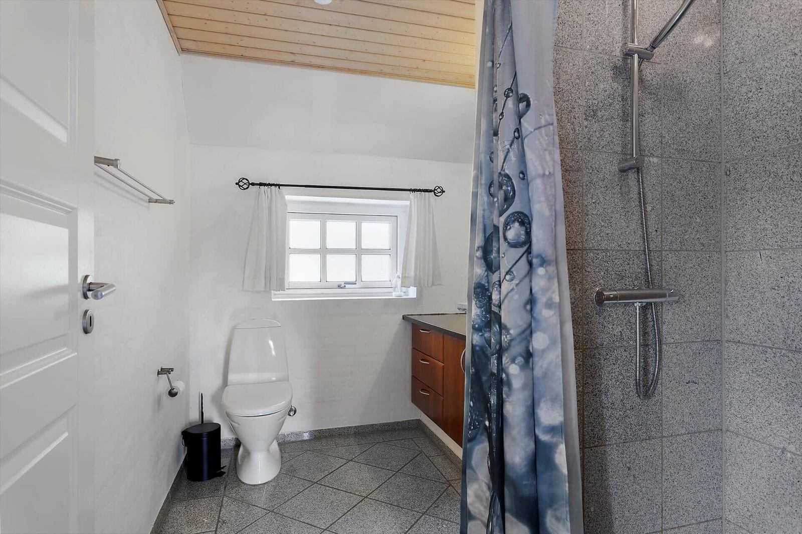 Badezimmer mit Dusche, Toilette und Fenster. Holzdecke und graue Fliesen.