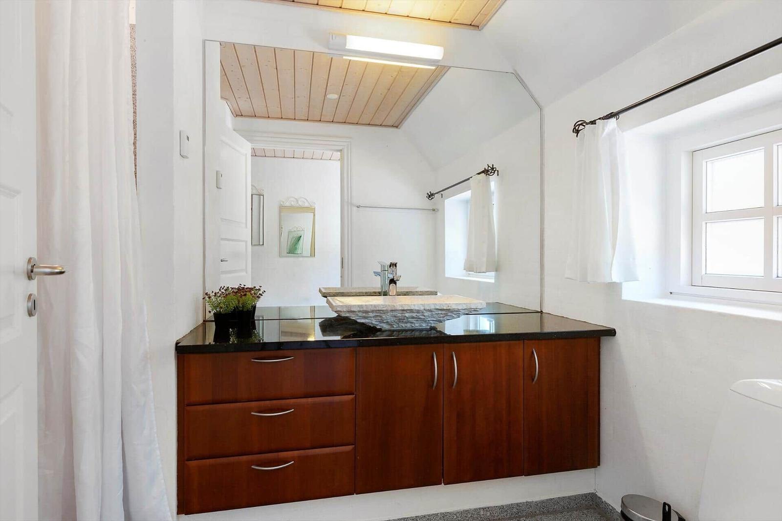 Badezimmer mit Holzschrank, Waschbecken und Spiegel. Fenster mit Vorhang.