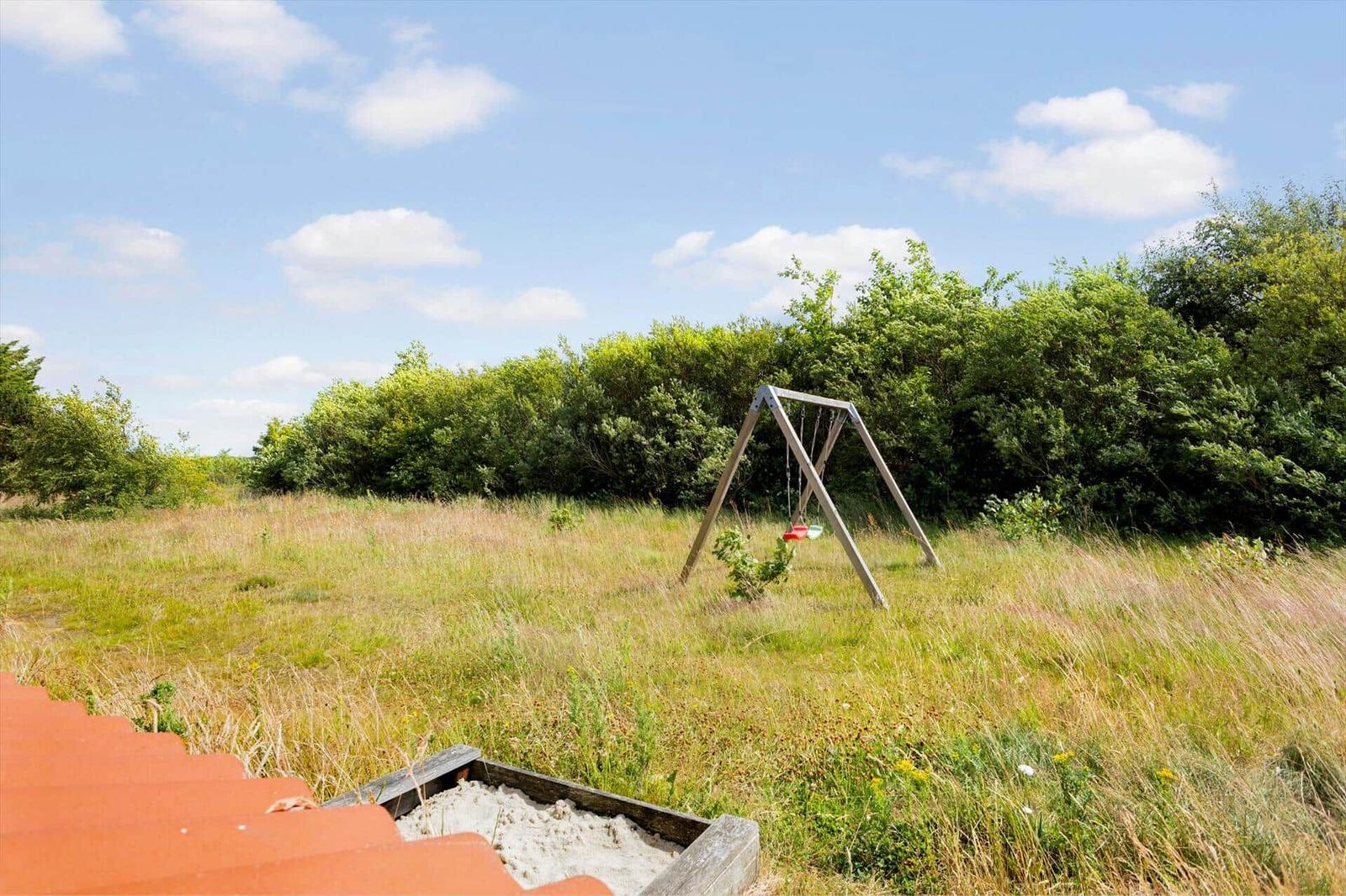 Ein Spielplatz mit Schaukel und Sandkasten im Gras. Hintergrund: dichter Busch und blauer Himmel.