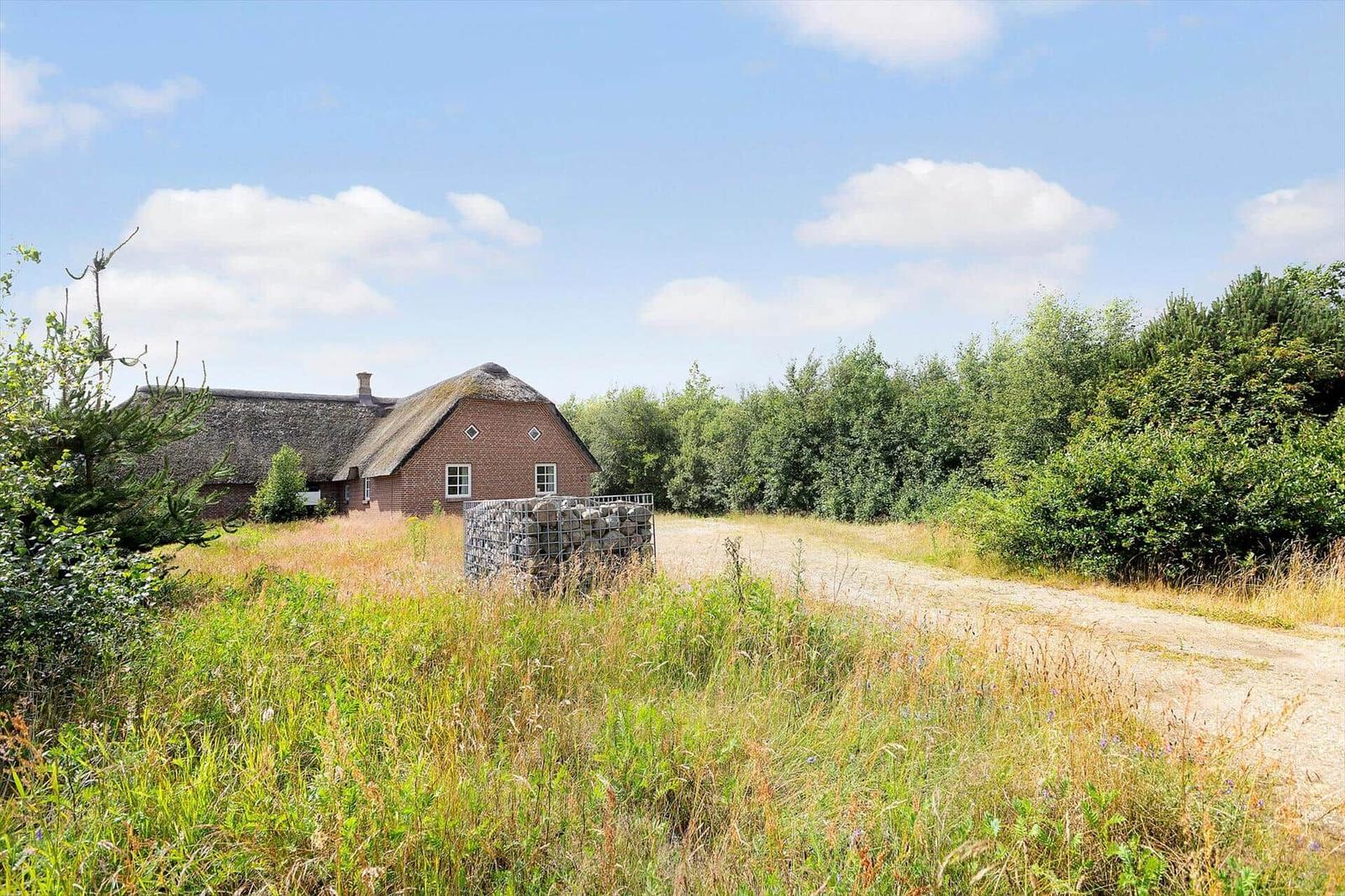 Rotes Backsteinhaus mit Strohdach, umgeben von Gras und Bäumen unter blauem Himmel.