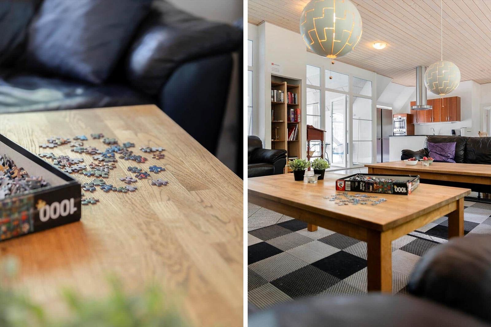 Ein Wohnzimmer mit Holztisch, Puzzle und schwarzen Ledersofas. Zwei runde Lampen hängen von der Decke.