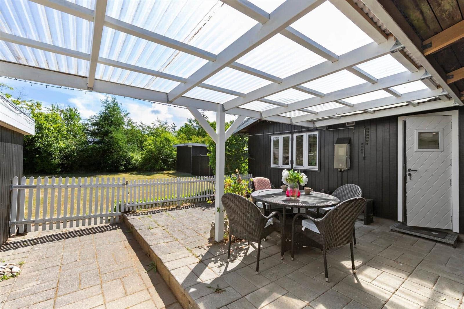 Terrasse mit Tisch und Stühlen unter weißem Dach. Hinterhof mit weißen Zaun und Bäumen.