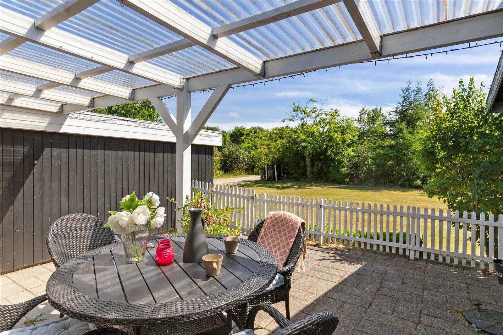 Terrasse mit Tisch, Stühlen und Blumen unter einem Dach. Hintergrund: weißer Zaun und Grünflächen.