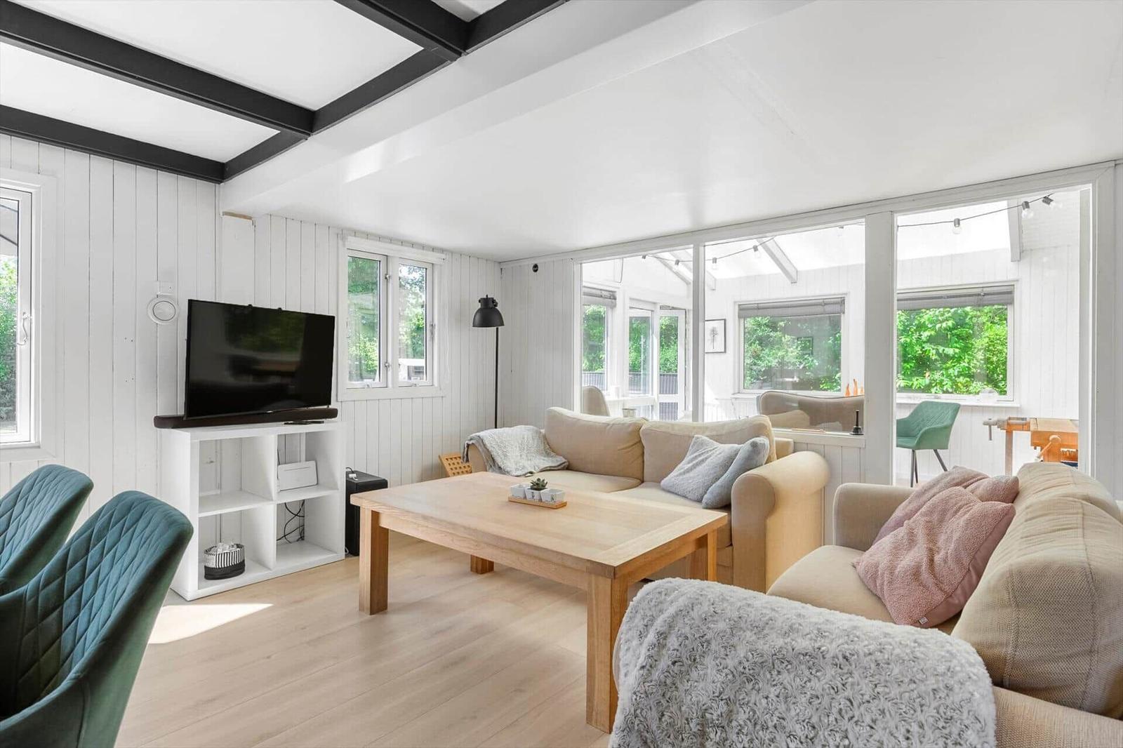 Wohnzimmer mit Sofa, Couchtisch und Fernseher. Große Fenster mit Blick auf Garten.