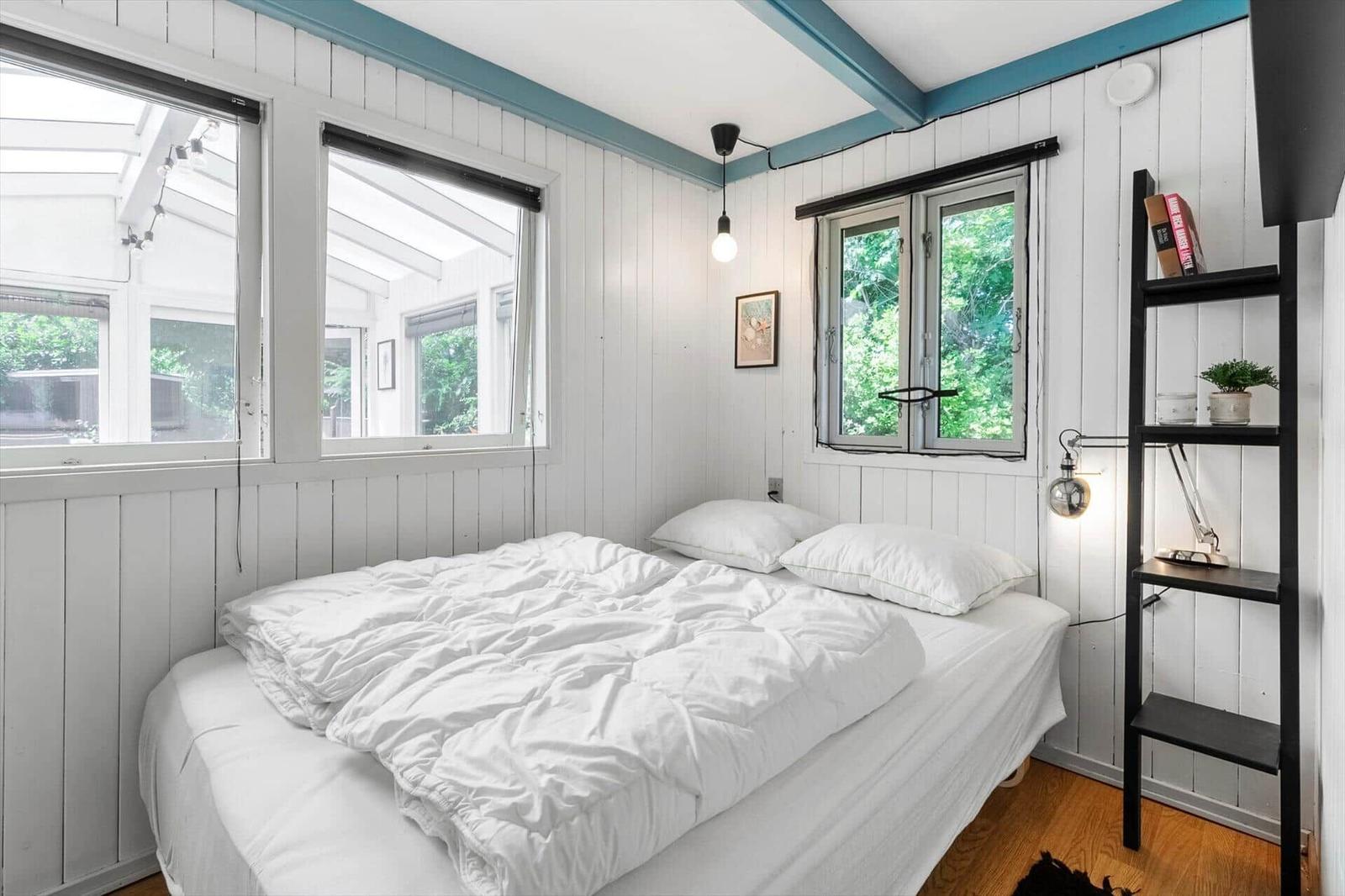 Schlafzimmer mit weißem Bett, Holzboden und Fenstern mit Blick auf Grün.