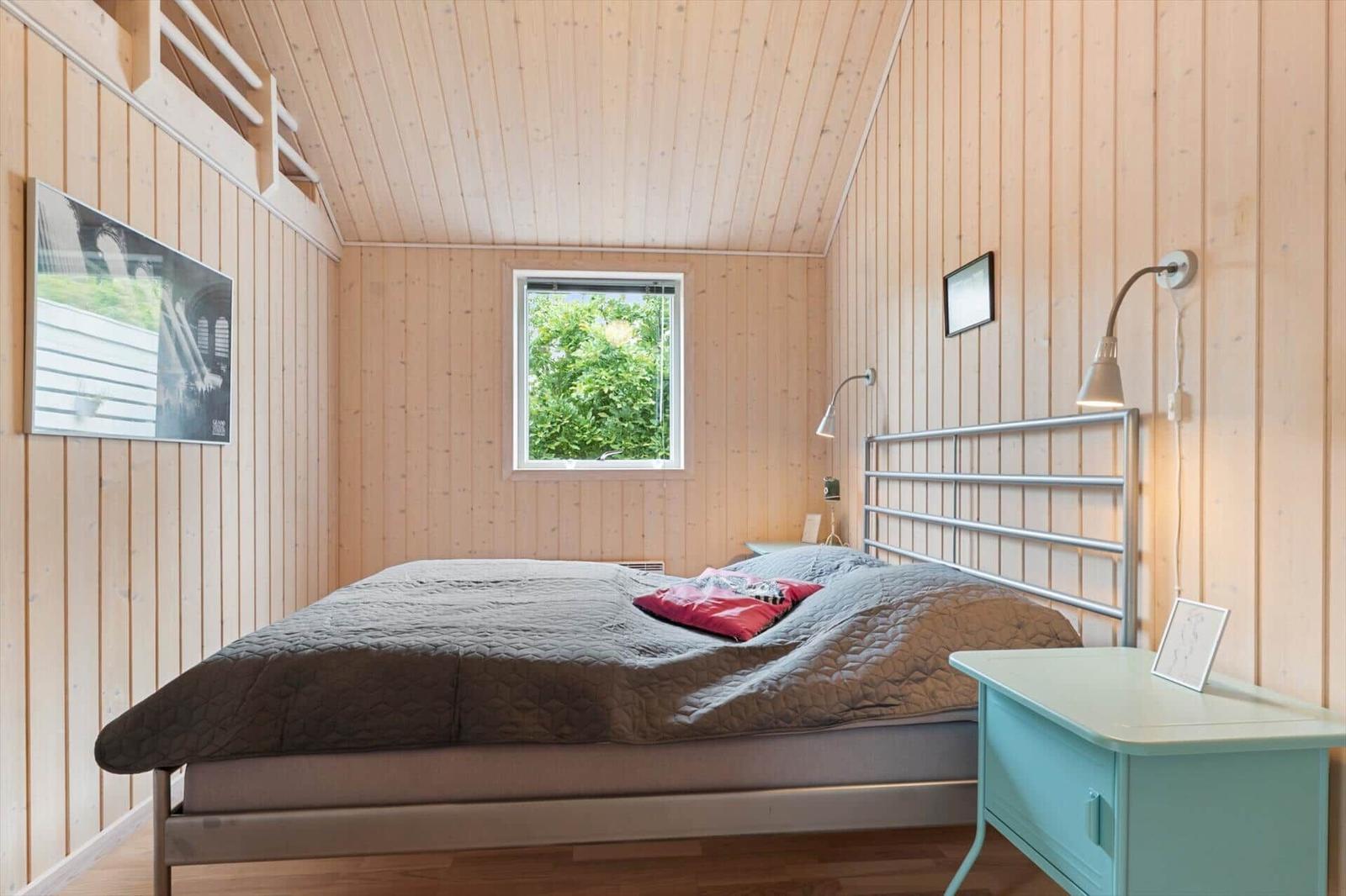 Schlafzimmer mit Holzwänden, Bett, Nachttisch und Fenster mit Blick auf Grün.