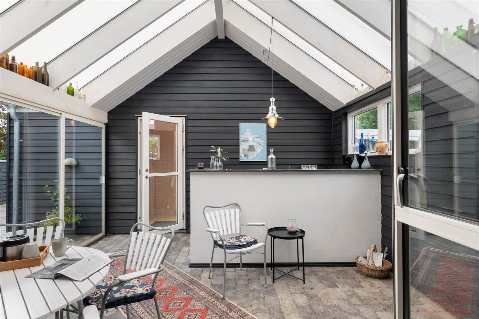 Terrasse mit Sitzgelegenheiten, Bar und Glasdach. Schwarze Holzwände und weiße Möbel.