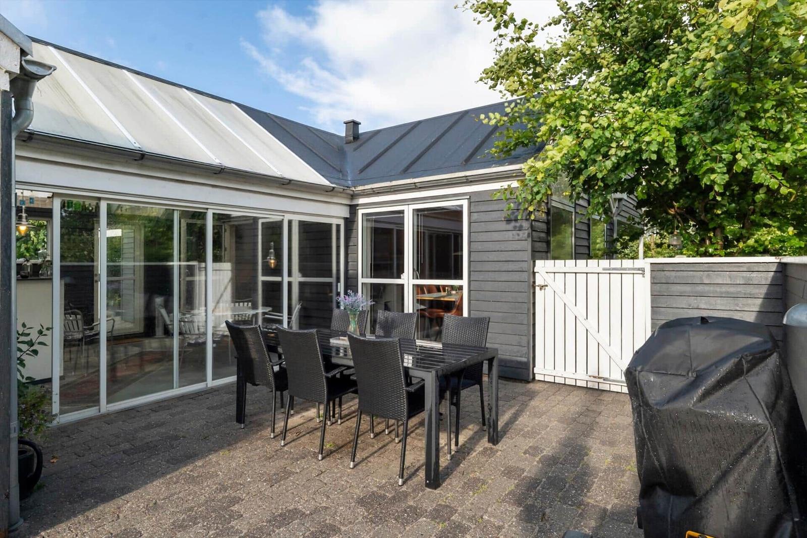 Terrasse mit Tisch und Stühlen, Glasveranda und Grillplatz. Hinterhof mit weißem Tor und Baum.