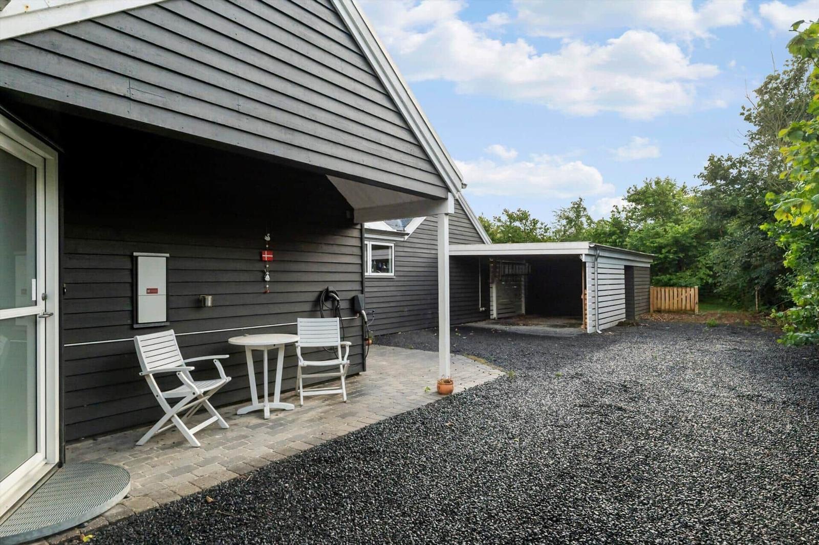 Haus mit Terrasse, weißem Tisch und Stühlen, Carport und Kiesfläche.