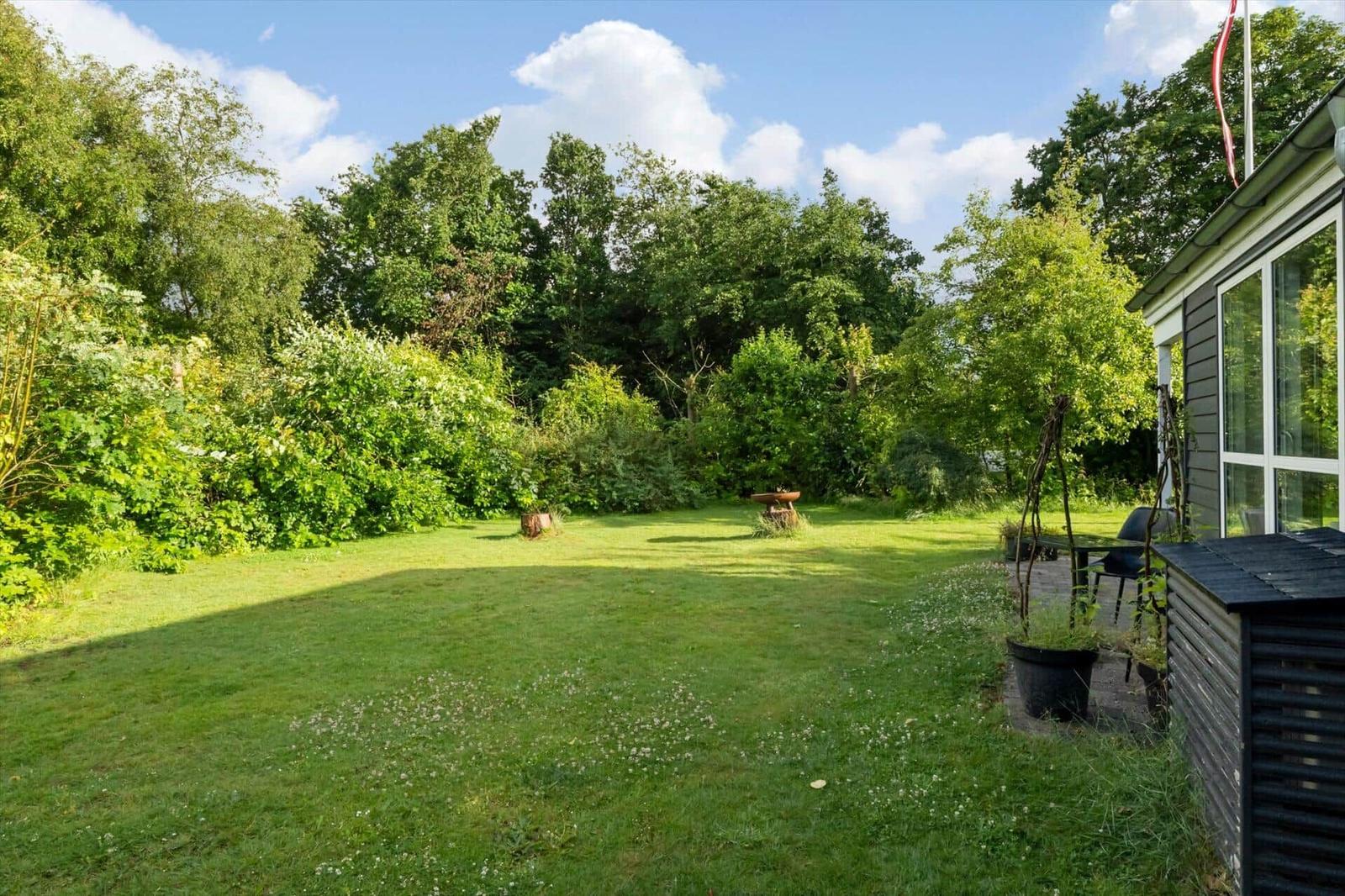 Grüner Garten mit Rasenfläche, Bäumen und einem Haus. Ein kleiner Tisch steht im Zentrum.
