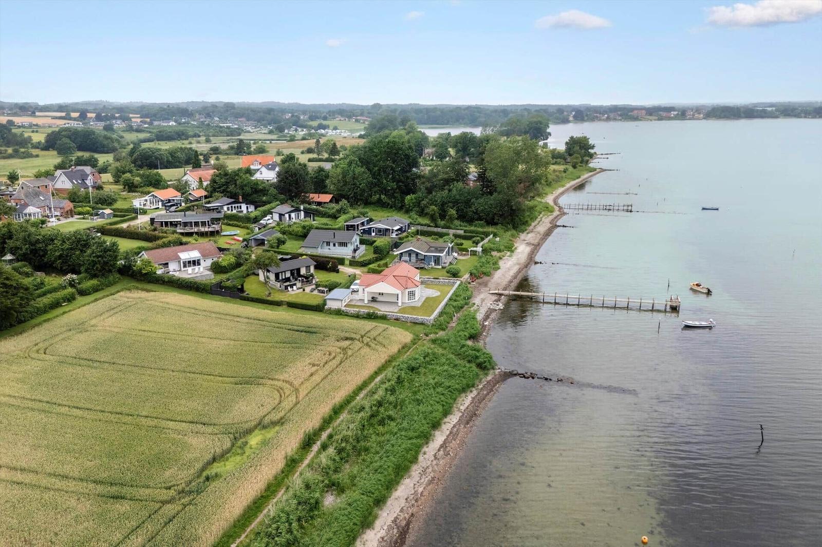Haus an ruhigem Seeufer mit Bootsanlegestelle und grünen Feldern im Hintergrund.