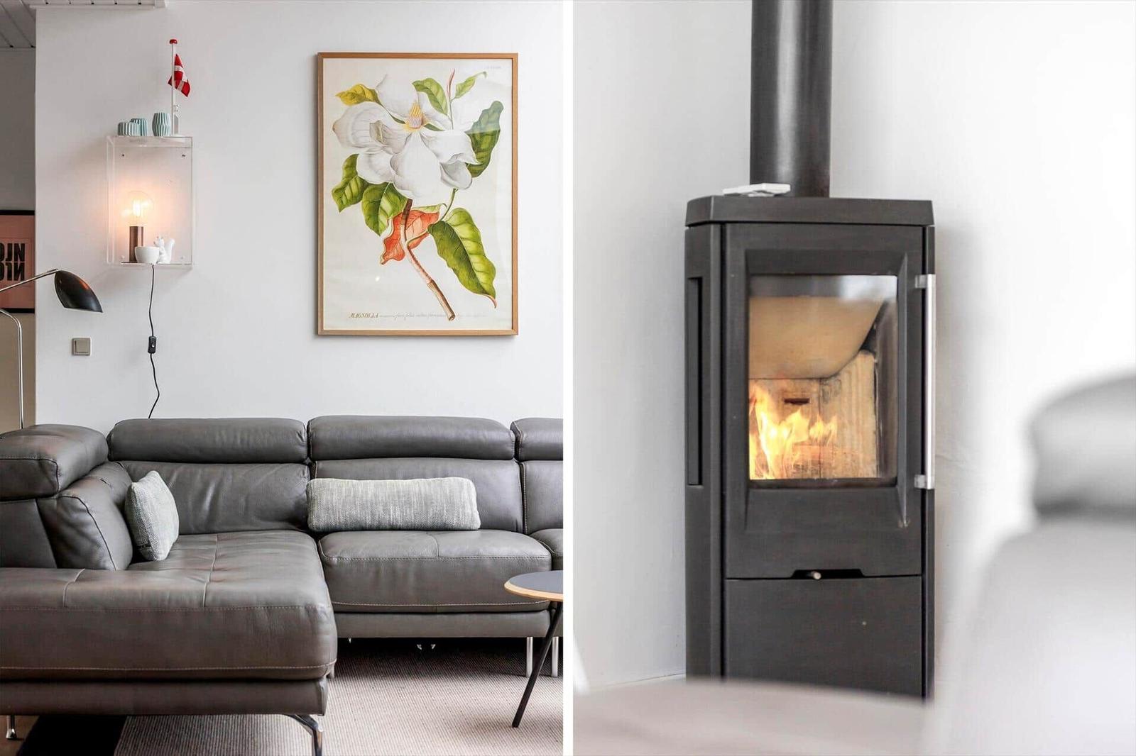 Graue Ledercouch mit Kissen unter einem Blumenbild. Holzofen mit brennendem Feuer.