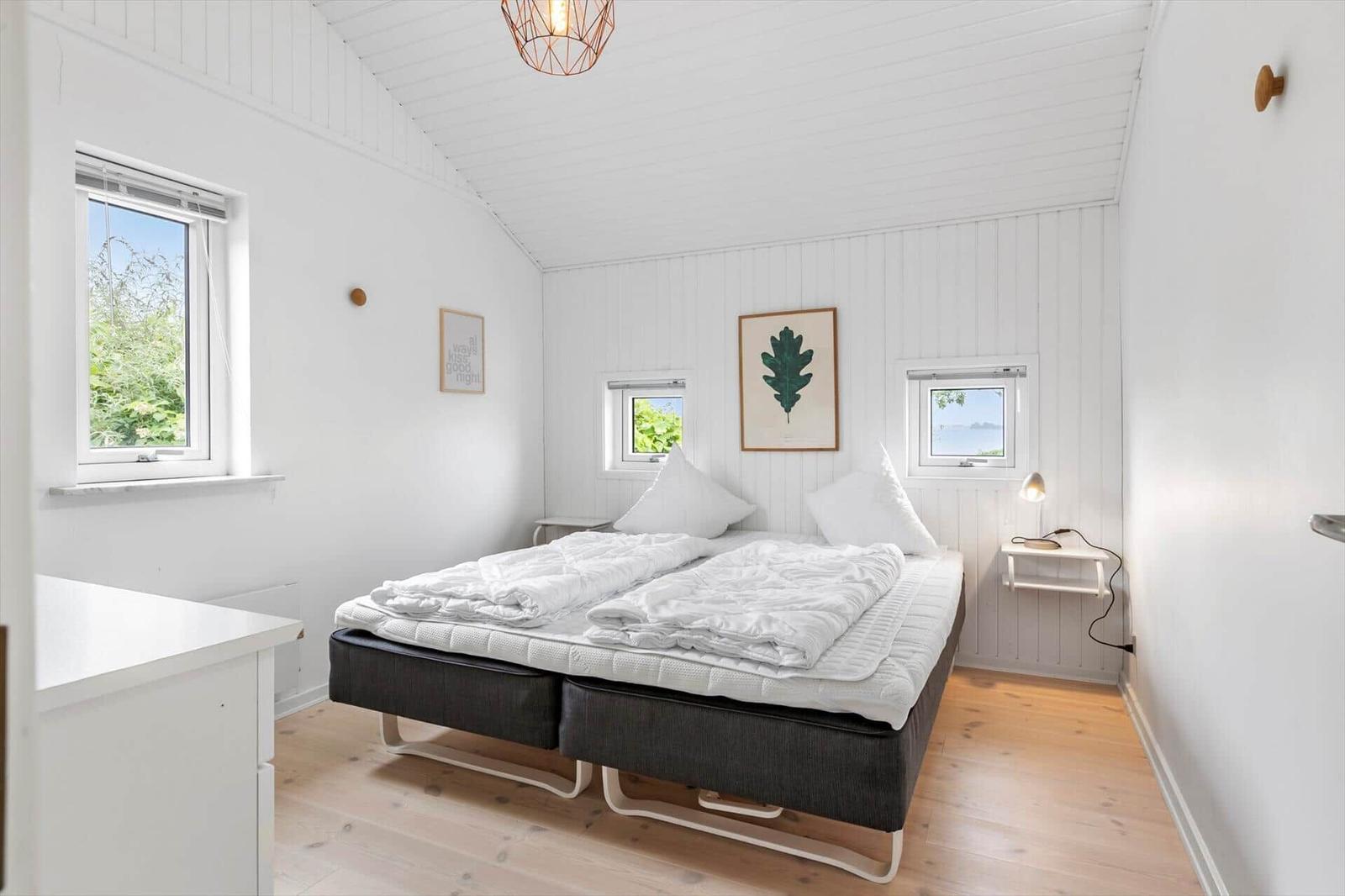 Schlafzimmer mit Bett, Fenstern und Wandbildern. Holzboden und weiße Wände.