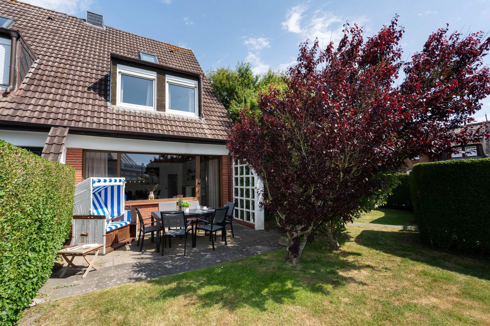Haus mit Terrasse, Garten und blau-weißem Liegestuhl. Roter Baum und grüne Hecke umgeben den Außenbereich.