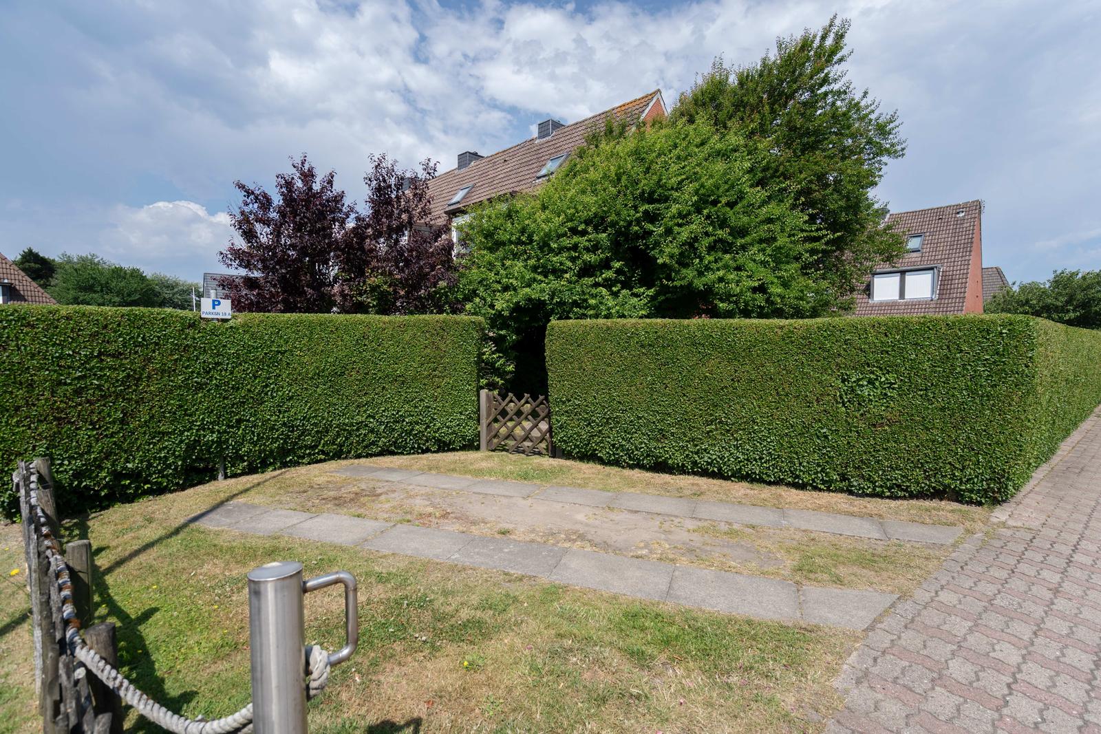 Garten mit Rasenfläche, gepflastertem Weg und hohem Heckenzaun. Im Hintergrund sind Bäume und ein Haus sichtbar.