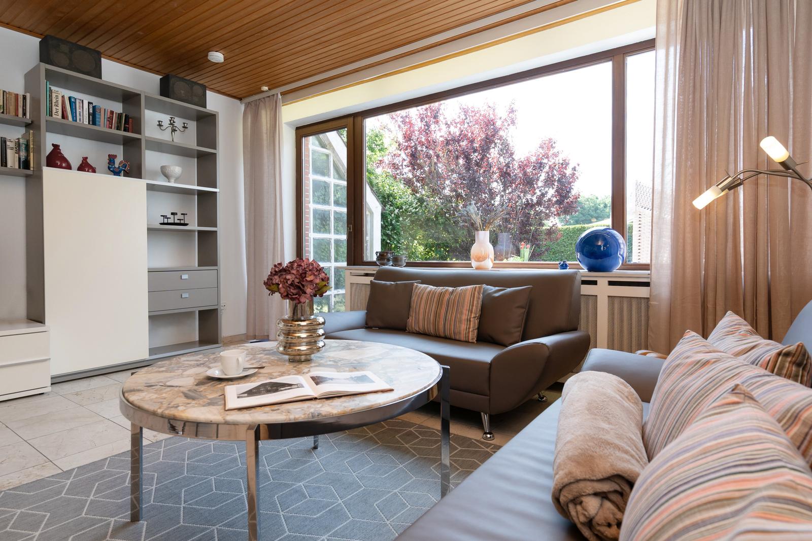 Wohnzimmer mit Sofa, runden Tisch und großen Fenstern. Blick auf Garten mit Bäumen.