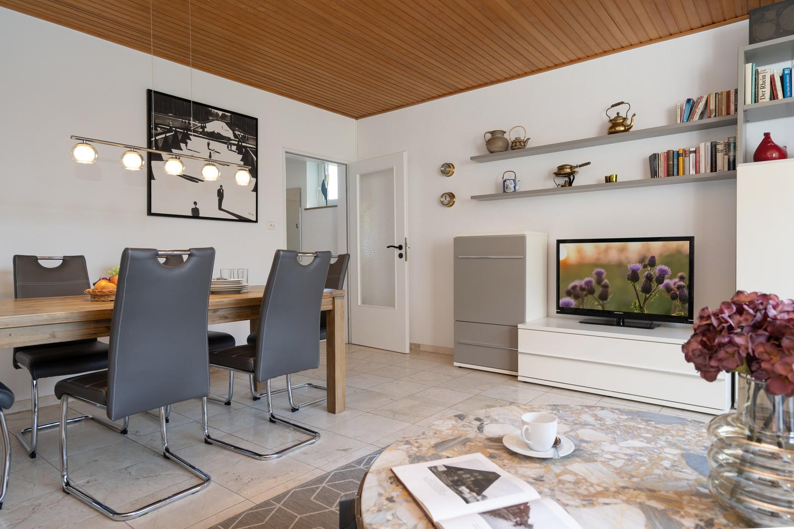 Essbereich mit Tisch und Stühlen, Wohnbereich mit Fernseher und Regalen.
