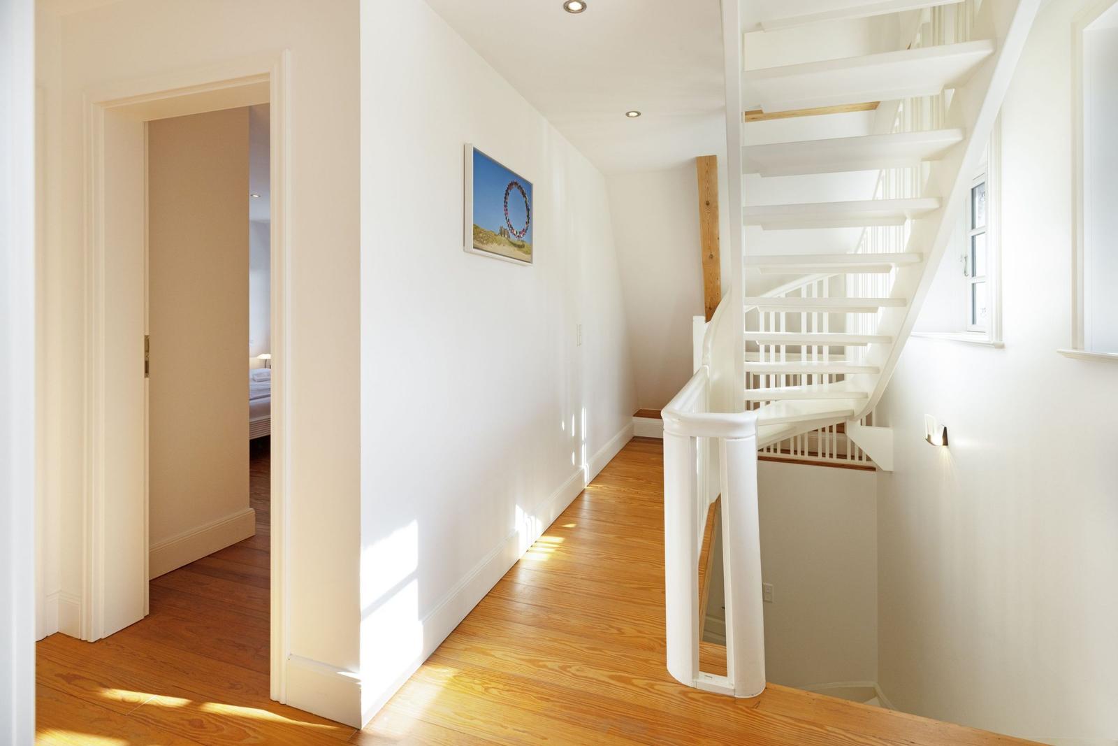 Flur mit Holzboden, weißer Treppe und Bild an der Wand. Tür führt in Schlafzimmer.