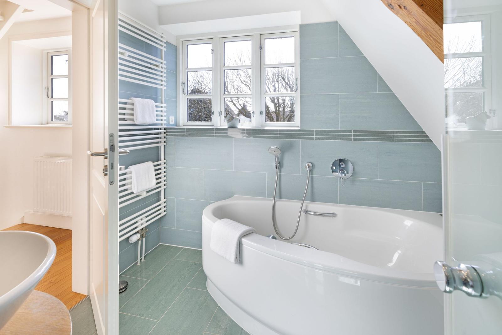 Bad mit Badewanne, Duschkopf und Fenster. Holzdielen und Heizkörper sichtbar.