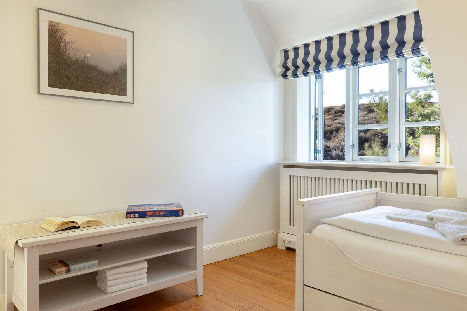 Schlafzimmer mit weißem Bett, Fenster mit blau-weißem Rollladen und Holzboden.