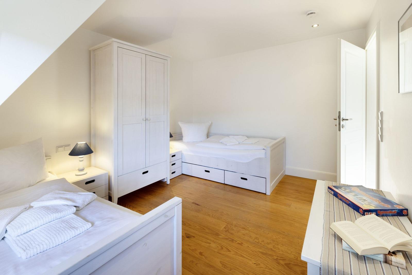 Zimmer mit zwei Betten, Schrank und Holzboden. Tisch mit Buch und Spiel.