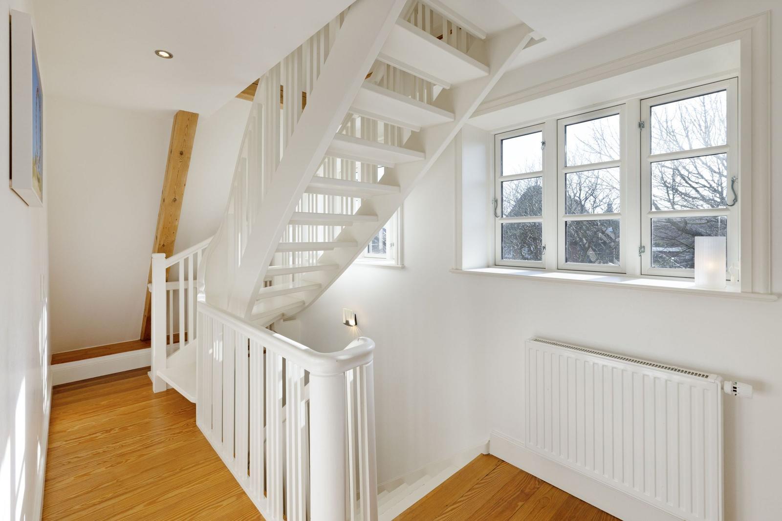 Helle Treppe mit weißem Geländer führt zur oberen Etage. Holzfußboden und Fenster mit Blick nach draußen.