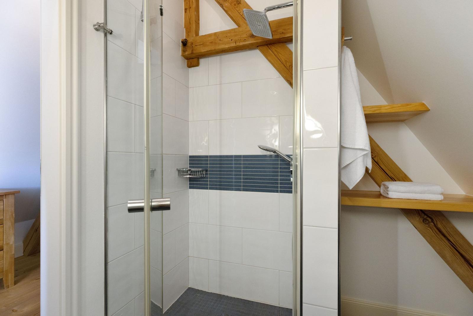 Dusche mit Glaswand, Holzbalken und Handtuchregal an geneigter Wand.