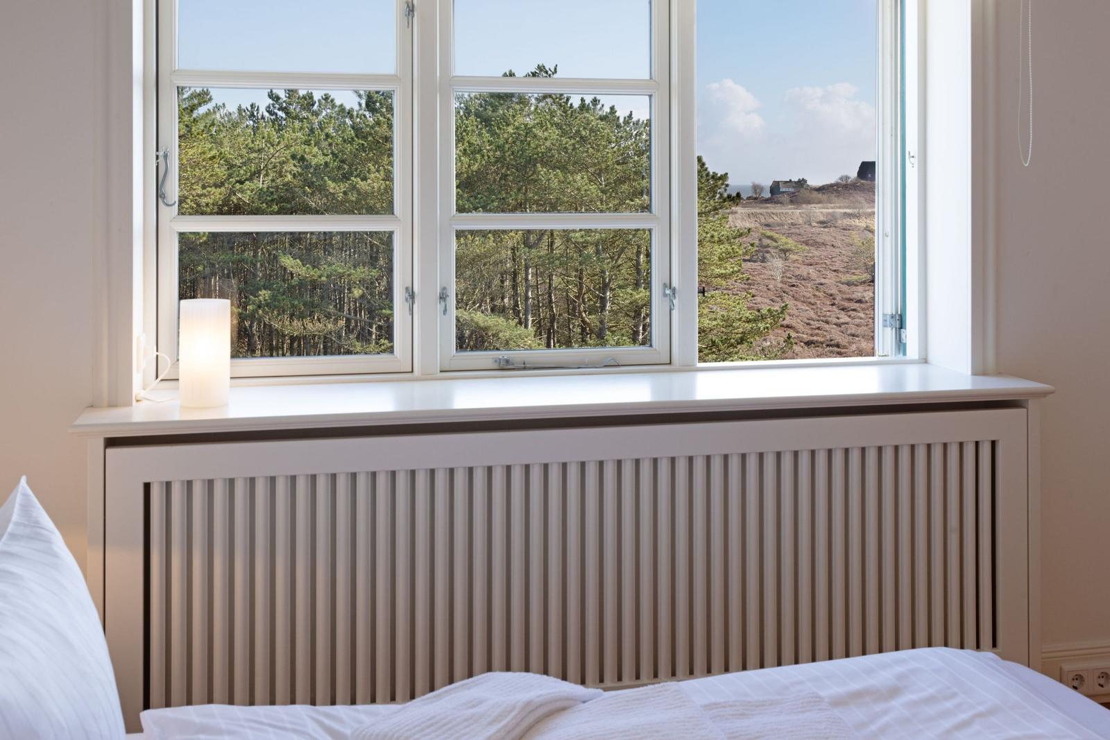 Ein Bett mit weißer Bettwäsche vor einem Fenster mit Blick auf Wald und Hügel.