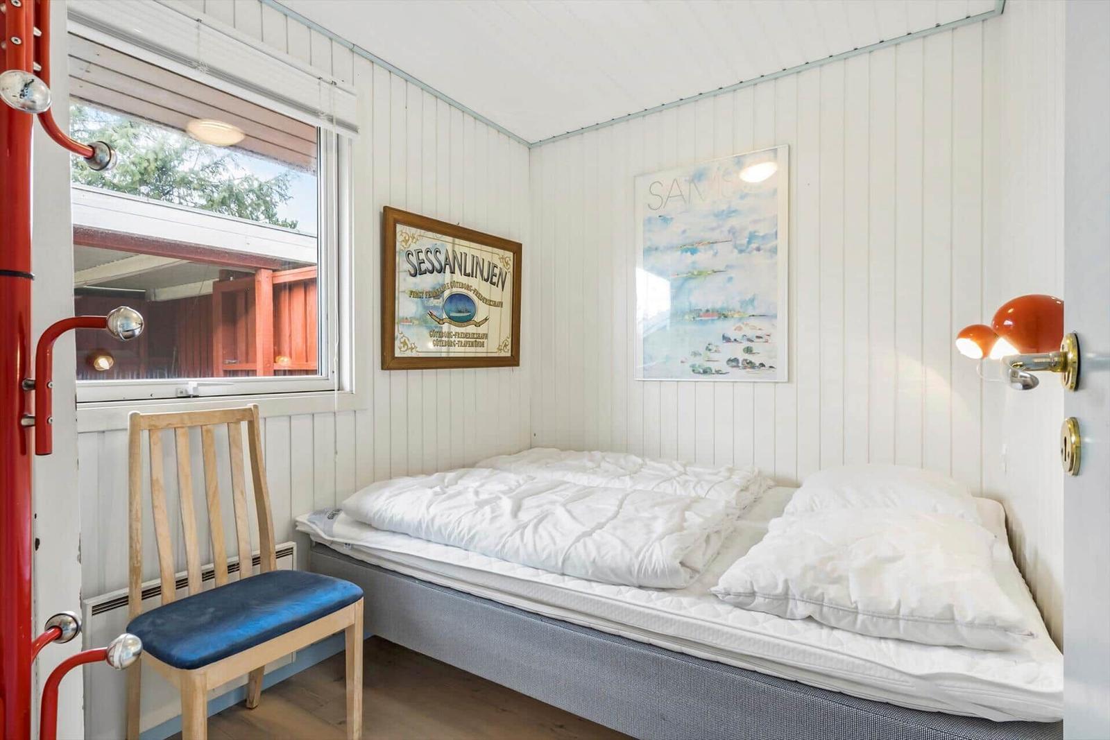 Schlafzimmer mit Bett, Stuhl und Fenster. Wände weiß verkleidet. Wandbilder und Nachttischlampen.